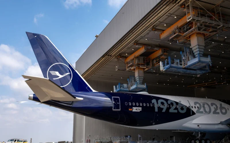 Lufthansa aircraft tail with logo near a large maintenance hangar and overhead equipment