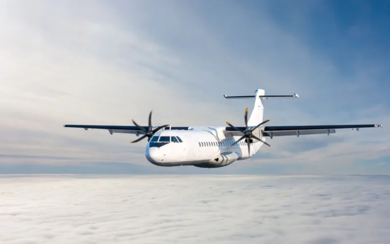White twin engine propeller airplane flying above a sea of clouds against a blue sky viewed from the front