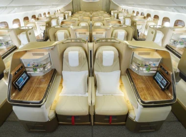 Luxurious business-class airplane cabin with beige lie-flat seats in private pods and wood-topped desks along a central aisle.