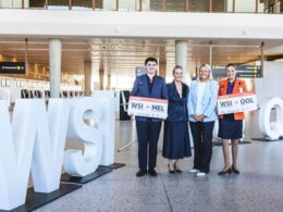 Four airline staff posing with oversized boarding pass signs in a modern airport terminal beside large white letters visible in the foreground