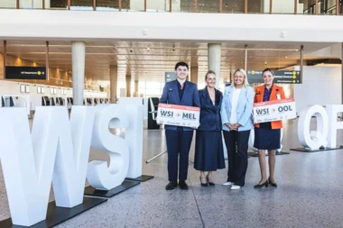Four airline staff posing with oversized boarding pass signs in a modern airport terminal beside large white letters visible in the foreground