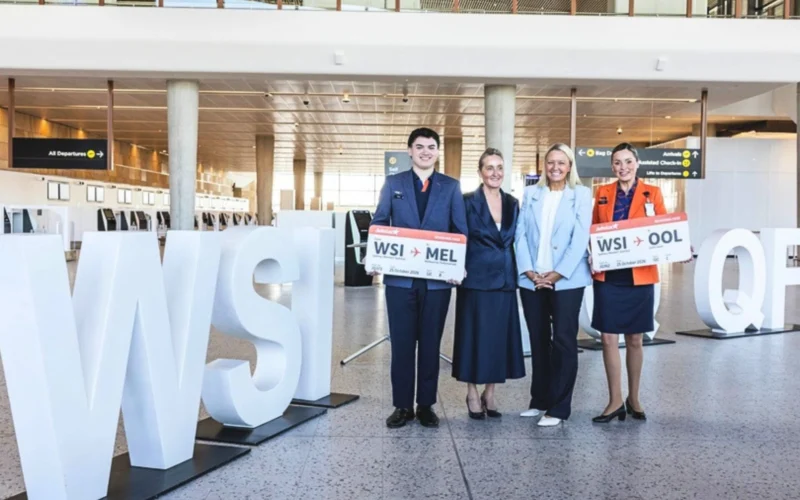 Four airline staff posing with oversized boarding pass signs in a modern airport terminal beside large white letters visible in the foreground