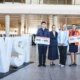 Four airline staff posing with oversized boarding pass signs in a modern airport terminal beside large white letters visible in the foreground