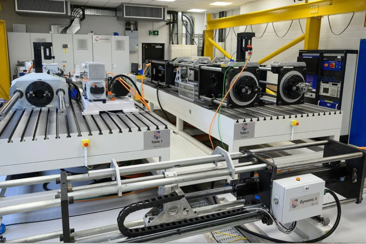 Collins Aerospace opens UK center for next gen aircraft tech Industrial lab with a long white testing bench and multiple motorized stages labeled Table 1 and Table 2 with cables and a yellow overhead crane in the background