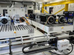 Industrial lab with a long white testing bench and multiple motorized stages labeled Table 1 and Table 2 with cables and a yellow overhead crane in the background