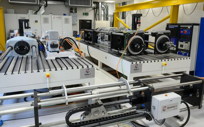 Industrial lab with a long white testing bench and multiple motorized stages labeled Table 1 and Table 2 with cables and a yellow overhead crane in the background
