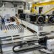 Industrial lab with a long white testing bench and multiple motorized stages labeled Table 1 and Table 2 with cables and a yellow overhead crane in the background