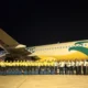 Night time group photo of airline staff in yellow shirts standing in front of a colorful commercial airplane on the tarmac