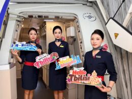 Three flight attendants in navy uniforms stand in an airplane doorway holding colorful welcome signs Hello Tbilisi Hello Shanghai