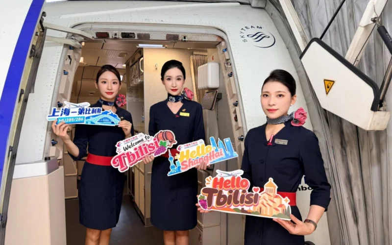 Three flight attendants in navy uniforms stand in an airplane doorway holding colorful welcome signs Hello Tbilisi Hello Shanghai