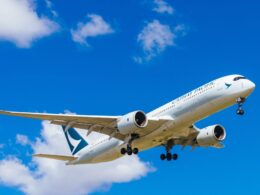 Cathay Pacific airliner in white livery ascending with its landing gear extended against a bright blue sky and scattered clouds