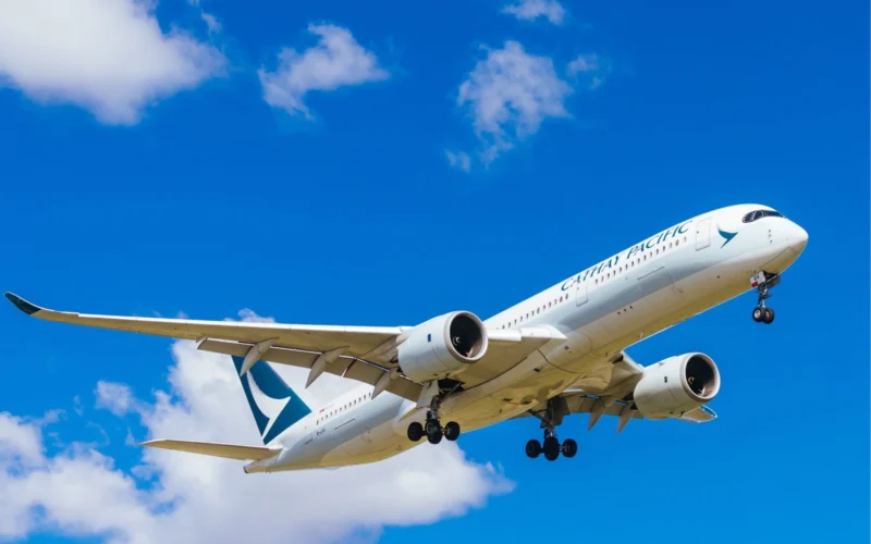 Cathay Pacific airliner in white livery ascending with its landing gear extended against a bright blue sky and scattered clouds