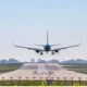 Front view of a blue passenger jet landing on a runway with a city skyline in the distance