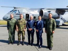 Five men stand in front of a Czech Air Force transport aircraft on a tarmac three are in olive green flight suits and two in business suits