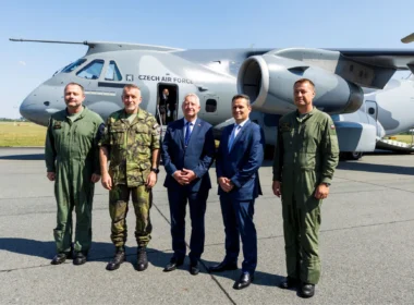 Five men stand in front of a Czech Air Force transport aircraft on a tarmac; three are in olive-green flight suits and two in business suits.