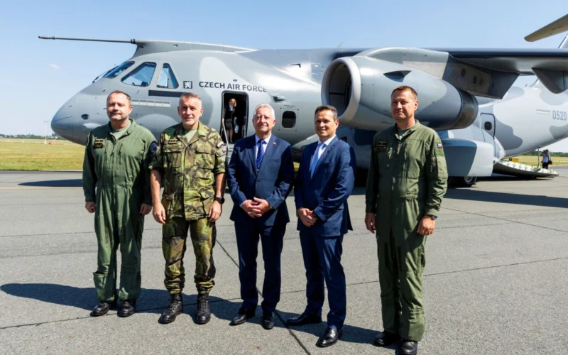 Five men stand in front of a Czech Air Force transport aircraft on a tarmac three are in olive green flight suits and two in business suits