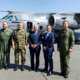 Five men stand in front of a Czech Air Force transport aircraft on a tarmac three are in olive green flight suits and two in business suits
