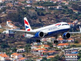British Airways airplane in landing attitude over hillside town with orange roofed houses and greenery behind