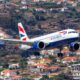 British Airways airplane in landing attitude over hillside town with orange roofed houses and greenery behind