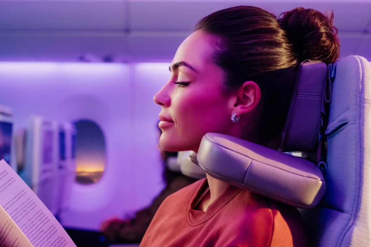 Emirates introduces U Dream headrest for economy class Female passenger on a plane resting with a neck pillow eyes closed reading a paper under purple cabin lighting
