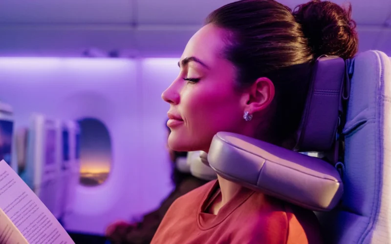 Female passenger on a plane resting with a neck pillow eyes closed reading a paper under purple cabin lighting