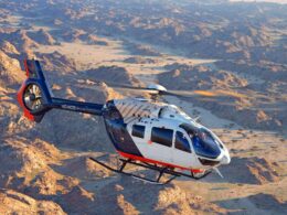 Blue and white Airbus helicopter in flight over a rugged desert canyon with rocky hills and dry riverbeds visible below  but keep concise
