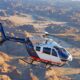 Blue and white Airbus helicopter in flight over a rugged desert canyon with rocky hills and dry riverbeds visible below  but keep concise