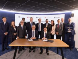 Group of executives in suits posing around a table as two leaders sign documents during a formal ceremony in front of a branded backdrop