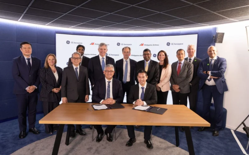 Group of executives in suits posing around a table as two leaders sign documents during a formal ceremony in front of a branded backdrop