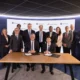 Group of executives in suits posing around a table as two leaders sign documents during a formal ceremony in front of a branded backdrop
