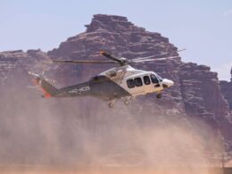 Passenger helicopter lifting off over a dusty desert with rocky cliffs in the background