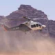 Passenger helicopter lifting off over a dusty desert with rocky cliffs in the background