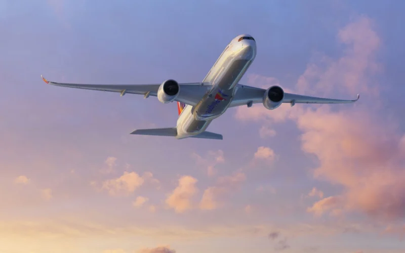 Commercial airliner photographed from below as it soars through a pastel sunset sky engines visible under the wings with a colorful tail