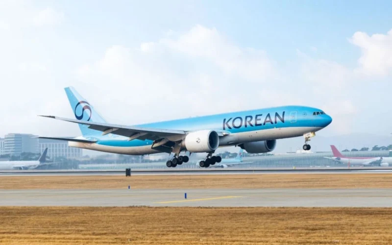 Korean Air jet lifting off from a runway with blue livery and tail logo visible against a clear sky airport in the background