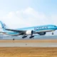 Korean Air jet lifting off from a runway with blue livery and tail logo visible against a clear sky airport in the background
