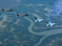 Four stealth fighter jets fly in formation above a winding braided river and green forested valley