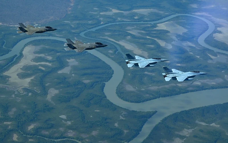 Four stealth fighter jets fly in formation above a winding braided river and green forested valley