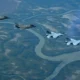 Four stealth fighter jets fly in formation above a winding braided river and green forested valley