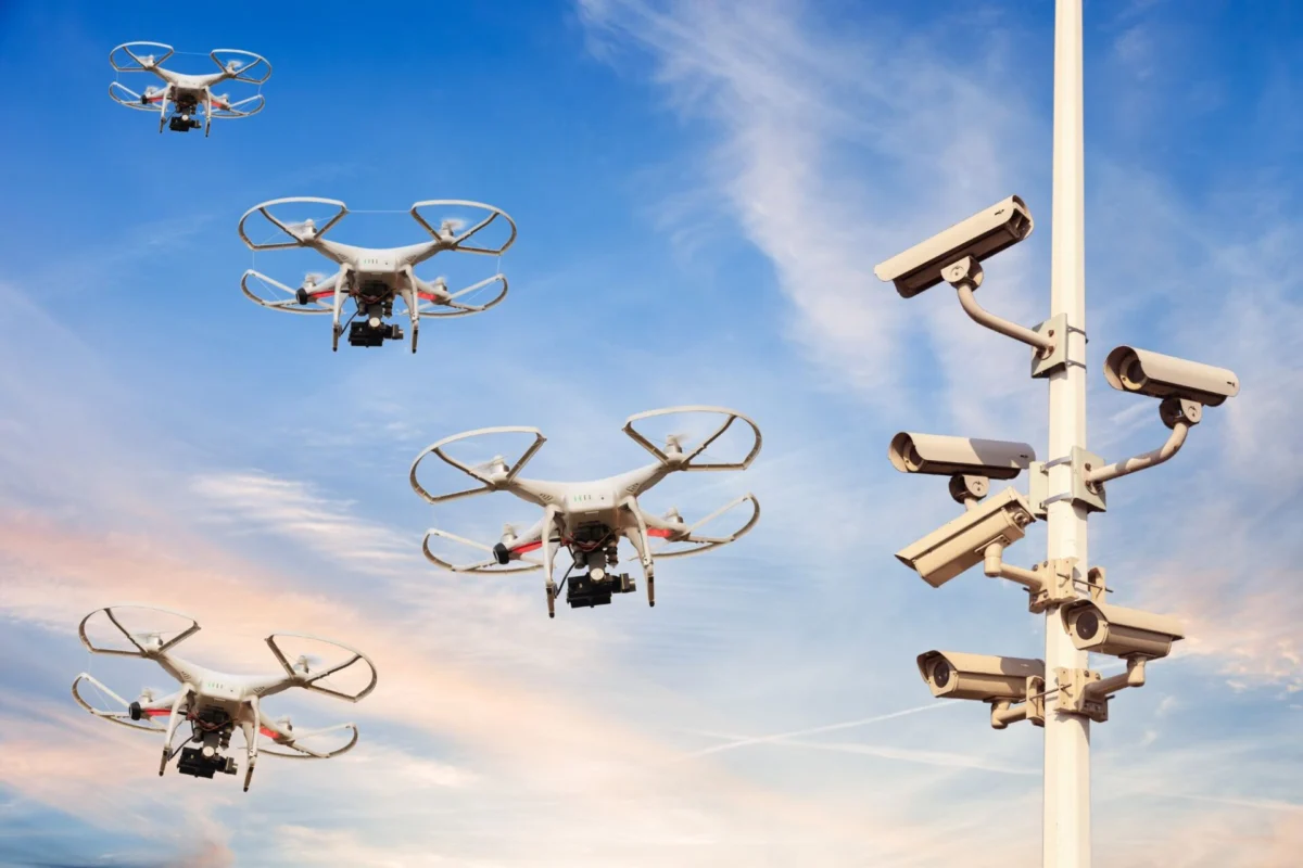 Quadcopter drones fly near a security camera tower against a blue sky at sunset