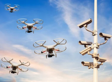 Quadcopter drones fly near a security camera tower against a blue sky at sunset.
