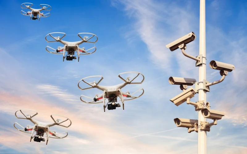 Quadcopter drones fly near a security camera tower against a blue sky at sunset