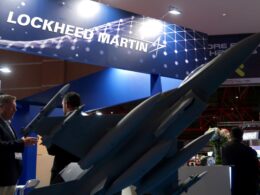 Professional trade show booth featuring Lockheed Martin branding with blue panels and aircraft models on display and people in suits discussing the exhibits
