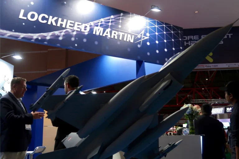 Professional trade show booth featuring Lockheed Martin branding with blue panels and aircraft models on display and people in suits discussing the exhibits