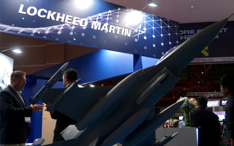 Professional trade show booth featuring Lockheed Martin branding with blue panels and aircraft models on display and people in suits discussing the exhibits