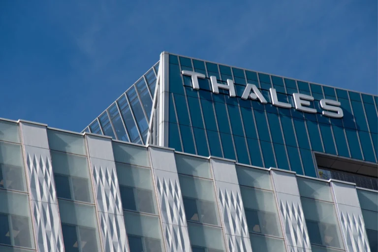 Modern glass office building with THALES signage on the roof against a clear blue sky