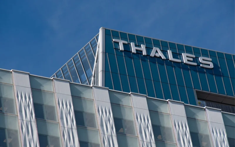 Modern glass office building with THALES signage on the roof against a clear blue sky