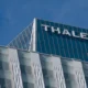 Modern glass office building with THALES signage on the roof against a clear blue sky