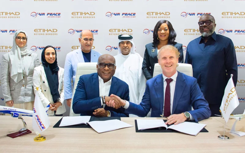 Two men in suits shake hands over signed documents at a conference table surrounded by colleagues with Air Peace and Etihad logos in the backdrop