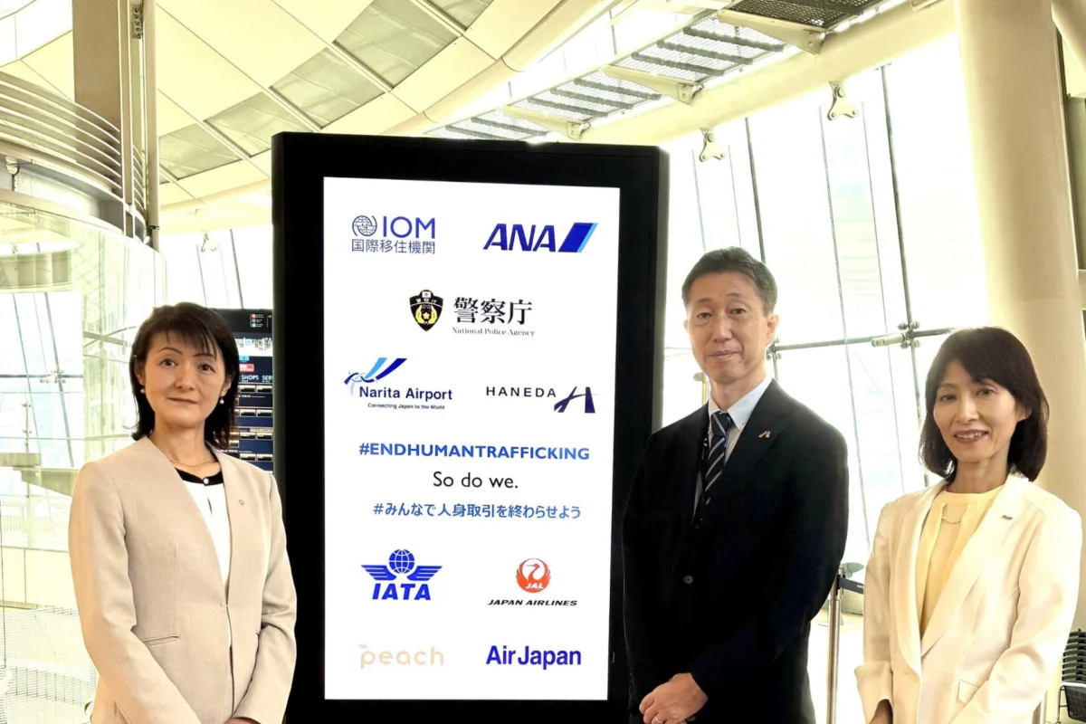 Three professionals in light colored suits posing beside a tall digital display board filled with airline logos and an anti trafficking message at an airport terminal