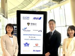 Three professionals in light colored suits posing beside a tall digital display board filled with airline logos and an anti trafficking message at an airport terminal